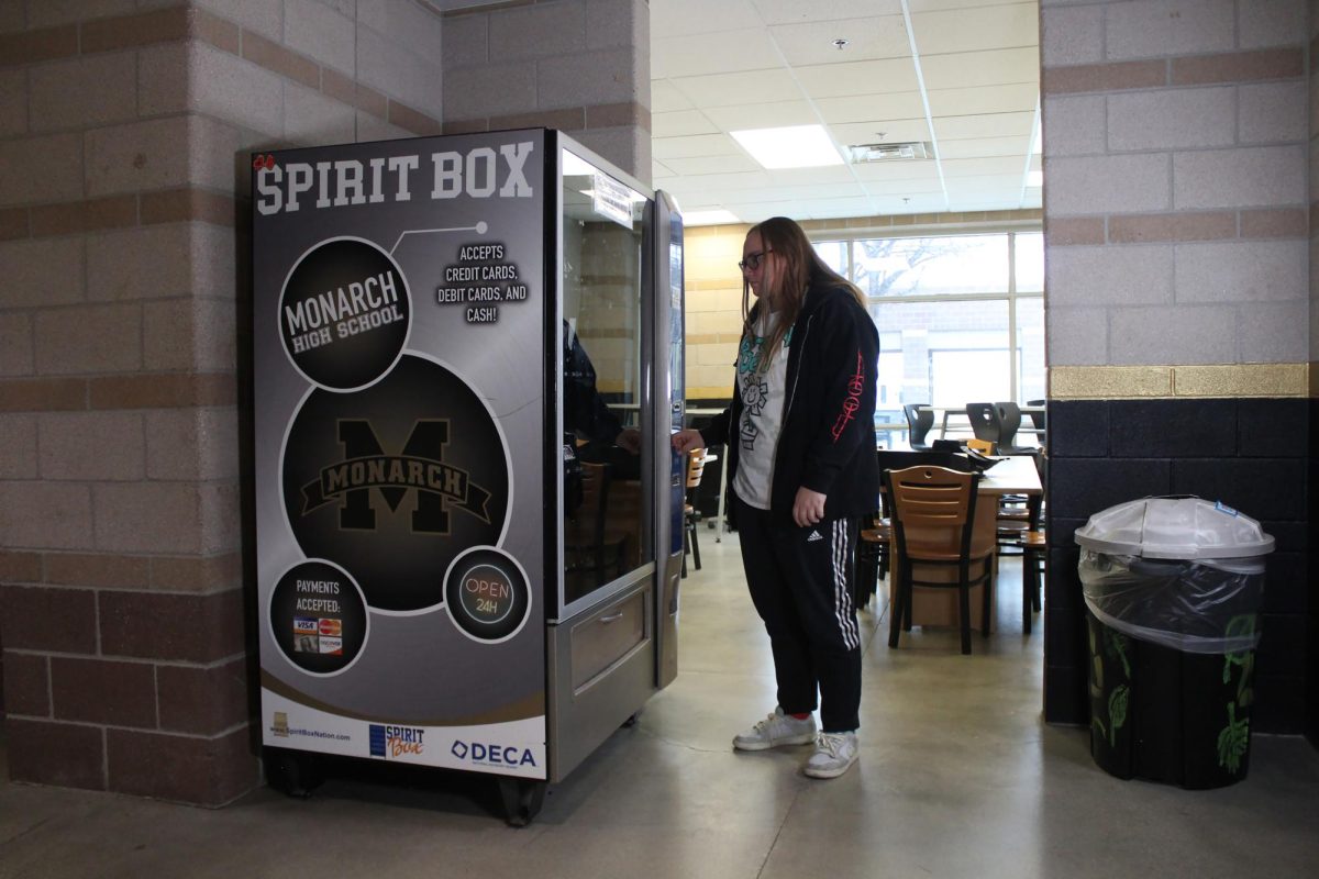 Standing in front of the DECA Spirit Box, Izabella Duckworth ‘25 awaits the reopening of the spirit wear vending machine. Scanning the different items in the machine, she sees  daily essentials and fun clothing items.